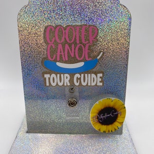 May include: A silver glitter badge holder with a blue and pink "Cooter Canoe Tour Guide" design. A sunflower sticker with the text "Wildflower Creations" is on the bottom right corner.