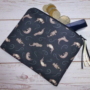 May include: A black zippered pouch with a repeating pattern of brown otters on a dark blue background. The pouch has a black zipper and a white and cream tassel.