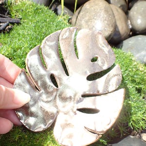 May include: A handcrafted, metallic Monstera leaf sculpture with a textured surface. The leaf features several cut-out holes and is held by a hand against a backdrop of green moss and stones. The sculpture has a brown and silver color.