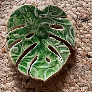 May include: A ceramic dish shaped like a monstera leaf with a glossy green crackle glaze finish. The surface features an embossed, swirling pattern throughout the leaf, with several cut-out sections characteristic of the plant's natural foliage.