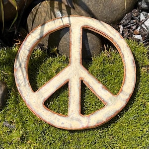 May include: A circular ceramic peace sign ornament featuring a cream-colored glaze with visible brown clay edges. The surface has a subtle, mottled texture throughout the circular frame and the interior intersecting lines of the peace symbol.
