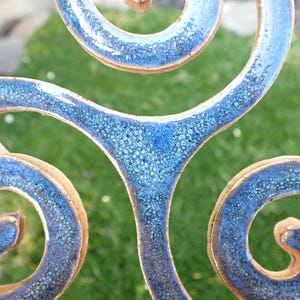 May include: A ceramic triskelion ornament featuring three interconnected spirals with a glossy, speckled blue glaze. The edges of the clay form are visible, showing a natural tan color that contrasts with the textured, deep blue finish of the surface.