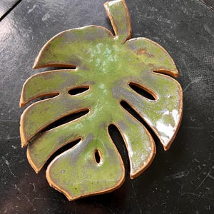 May include: A ceramic monstera leaf dish featuring a glossy, mottled green glaze with dark brown undertones. The leaf shape includes characteristic cutouts and a stem at the top, with a smooth, reflective surface and visible clay edges.