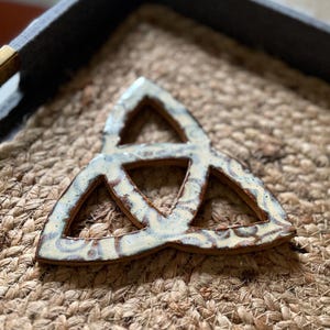 Può includere: Un ornamento in ceramica a forma di triquetra con una smaltatura color crema lucida caratterizzata da macchie marroni e motivi a vortice. Il design a nodo triangolare presenta tre sezioni aperte e una superficie liscia e riflettente.
