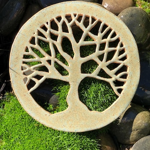 May include: A circular ceramic wall plaque featuring a cutout design of a tree with spreading branches and a thick trunk. The piece has a speckled, light tan and pale green glaze finish, with the intricate tree silhouette creating open spaces.