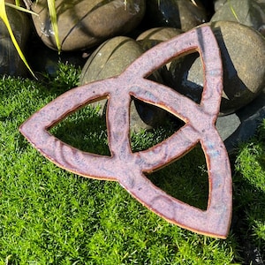 May include: A ceramic triquetra symbol featuring a glossy, mottled glaze in shades of dusty rose, lavender, and hints of blue. The triangular knotwork design has three interconnected loops with a smooth, reflective surface and defined edges.