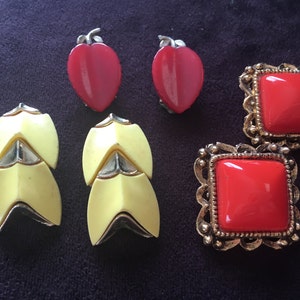 May include: Four pairs of vintage earrings. Two pairs are red and have a heart shape. One pair is yellow and has a geometric design. The other pair is red and has a square shape with a gold frame.