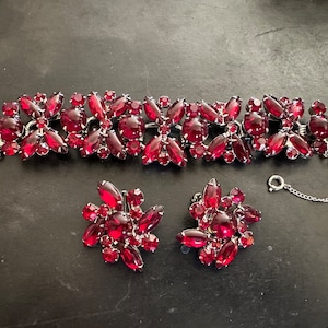 May include: A vintage red rhinestone bracelet and earrings set. The bracelet is made of silver tone metal and features a floral design with red rhinestones. The earrings are clip-on and match the bracelet.