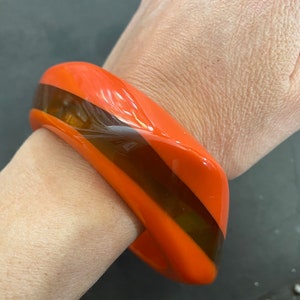 Op de afbeelding: Oranje en bruine plastic armband. De armband is dik en heeft een gebogen vorm.