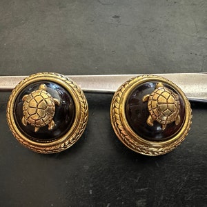 May include: A pair of gold-tone earrings with a black enamel turtle design. The turtles are detailed with gold accents and are set in a round, textured frame.