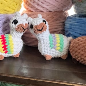 May include: Handmade crochet llamas with colorful accents and cactus plushies in red, green, and yellow. The llamas have rainbow-colored blankets. The background features various colors of yarn, suggesting a craft or home decor theme.