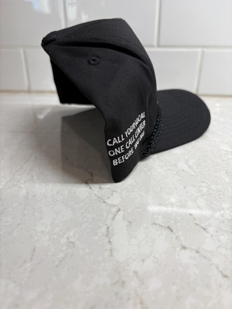 Pu&ograve; includere: Cappellino da baseball nero con testo ricamato bianco: "CALL YOUR LOCAL ONE CALL CENTER BEFORE YOU DIG". Il cappellino ha una visiera curva e un dettaglio intrecciato sul bordo.