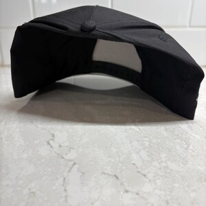 Pu&ograve; includere: Un cappellino da baseball nero con visiera curva. Il cappellino ha un bottone sulla parte superiore e una chiusura a scatto regolabile sul retro. L'interno &egrave; bianco. Il cappellino &egrave; appoggiato su una superficie bianca e grigia.
