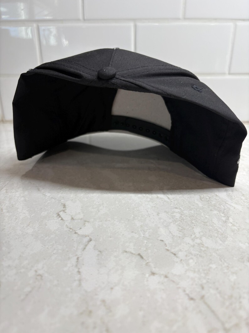 Pu&ograve; includere: Un cappellino da baseball nero con visiera curva. Il cappellino ha un bottone sulla parte superiore e una chiusura a scatto regolabile sul retro. L'interno &egrave; bianco. Il cappellino &egrave; appoggiato su una superficie bianca e grigia.
