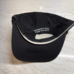 Pu&ograve; includere: Cappellino da baseball nero con visiera curva e chiusura regolabile a scatto. Il lato inferiore della visiera &egrave; bianco. Il testo "Emergency Only 1-888-367-6671" &egrave; ricamato sul cappellino.