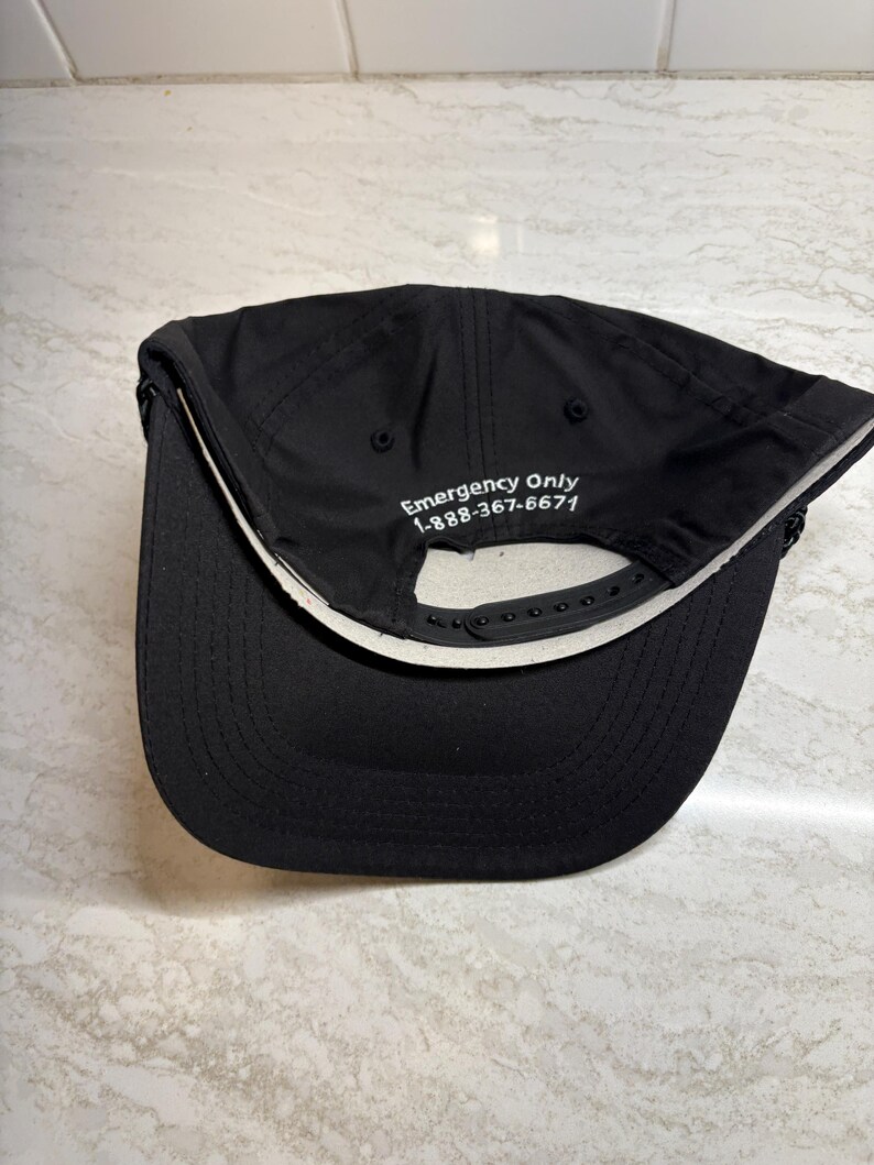 Pu&ograve; includere: Cappellino da baseball nero con visiera curva e chiusura regolabile a scatto. Il lato inferiore della visiera &egrave; bianco. Il testo "Emergency Only 1-888-367-6671" &egrave; ricamato sul cappellino.