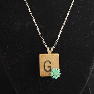 Puede incluir: Un collar de cadena de plata con un colgante que presenta una baldosa de madera cuadrada con la letra "G" y una flor turquesa.