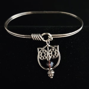May include: A silver bangle bracelet with a dangling owl charm. The owl is made of silver and has a dark purple crystal in its center.