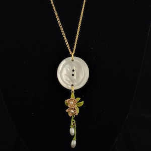 May include: A gold chain necklace with a white mother-of-pearl button pendant and a floral charm with pink and green accents.