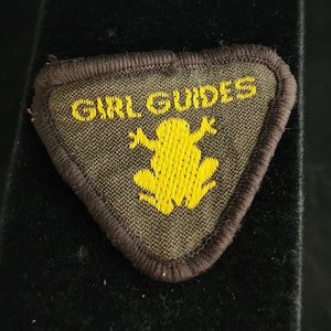 May include: A triangular Girl Guides patch with a brown border and background. The patch features the words "GIRL GUIDES" in yellow, along with a yellow frog emblem. The patch is likely made of fabric and is designed to be sewn onto clothing.