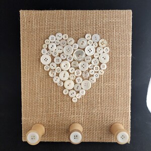 May include: A square burlap board with a heart made of white buttons. Three wooden pegs with white buttons are attached to the bottom. The background is black.