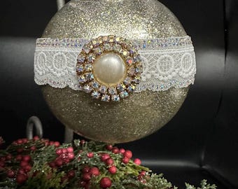 Può includere: Un ornamento natalizio dorato glitterato con una fascia di pizzo bianco e un abbellimento centrale tempestato di gioielli. L'ornamento ha un centro a forma di perla ed è sospeso su uno sfondo scuro. Decorazioni festive per le vacanze.