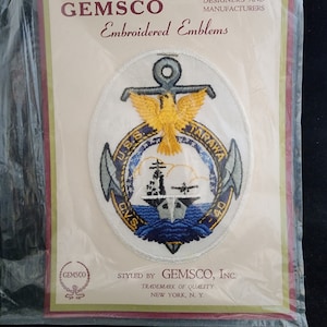Puede incluir: Un parche bordado blanco con un ancla azul, un águila amarilla y el texto "USS TARAWA CV-40". El parche está rodeado por un borde blanco y está en una tarjeta blanca con el texto "GEMSCO Embroidered Emblems" y "STYLED BY GEMSCO, INC. TRADEMARK OF QUALITY NEW YORK, N. Y."