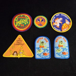 May include: A collection of nine embroidered patches featuring various themes, including a cookie festival, a space age roundup, a fall campout, and water fun. The patches are colourful and feature various designs, including stars, balloons, a rocket ship, a frog, and a camp scene.