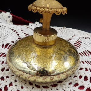 May include: A round, gold-toned glass perfume bottle with a textured, crackled metallic finish. The bottle features a gold-colored metal neck and a decorative, circular gold cap with a scalloped, filigree-style edge and a central textured dome.