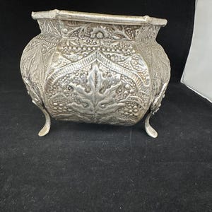 Puede incluir: Un macetero de plata con un intrincado diseño floral y tres patas. El macetero está hecho de metal y tiene una base cuadrada.