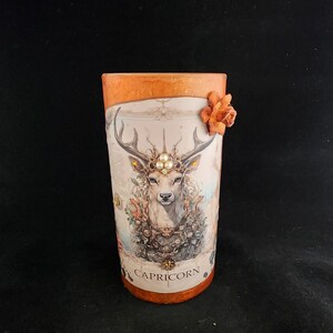 May include: A white ceramic candle holder with an orange top and a floral design. The candle holder features a deer with antlers and a crown, and the text "CAPRICORN" at the bottom.