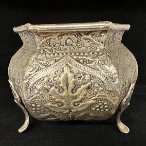 May include: A silver planter with an ornate design featuring leaves and grapes. The planter has a square top and three legs.
