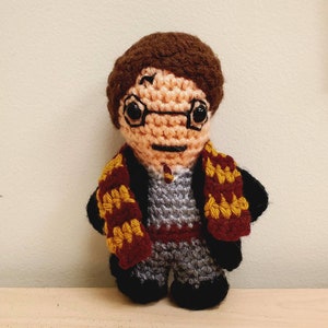 May include: A crocheted amigurumi figure of a young boy with brown hair, wearing a grey sweater, black pants, and a red and gold scarf.