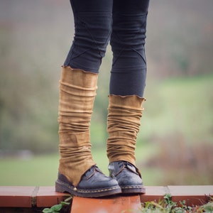 May include: A pair of brown leather leg warmers worn over black trousers and black boots. The leg warmers are gathered at the top and bottom, creating a pleated look.