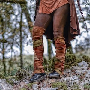 Op de afbeelding: Een paar bruine leren leggings met kleurrijke patchwork beenwarmers. De leggings en beenwarmers worden gedragen door een persoon die op een rotsachtig oppervlak staat met bomen op de achtergrond.