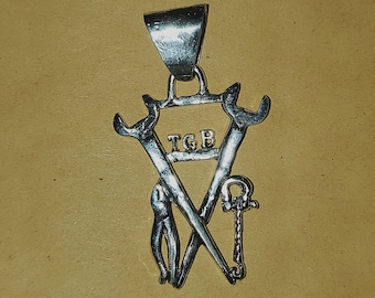 Op de afbeelding: Zilveren hanger met een driehoekig ontwerp met twee gekruiste moersleutels en een sleutel. De hanger heeft de letters "TGB" in het driehoekige ontwerp.