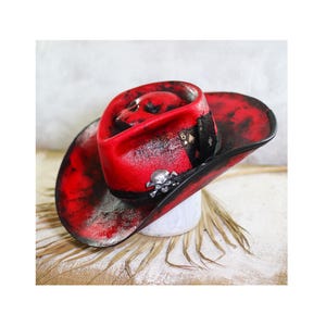 Könnte beinhalten: Ein roter Cowboyhut mit schwarzer und weißer Sprühfarbe und einem Totenkopfdesign. Der Hut hat eine breite Krempe und eine hohe Krone.