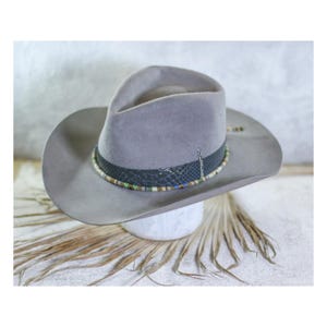 May include: A grey felt cowboy hat with a black leather band and a beaded trim. The hat is sitting on a light brown surface.
