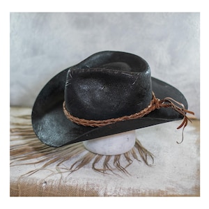 May include: A black cowboy hat with a wide brim and a braided brown leather band. The hat is displayed on a white pedestal, with a neutral background. The hat has a distressed, textured appearance.