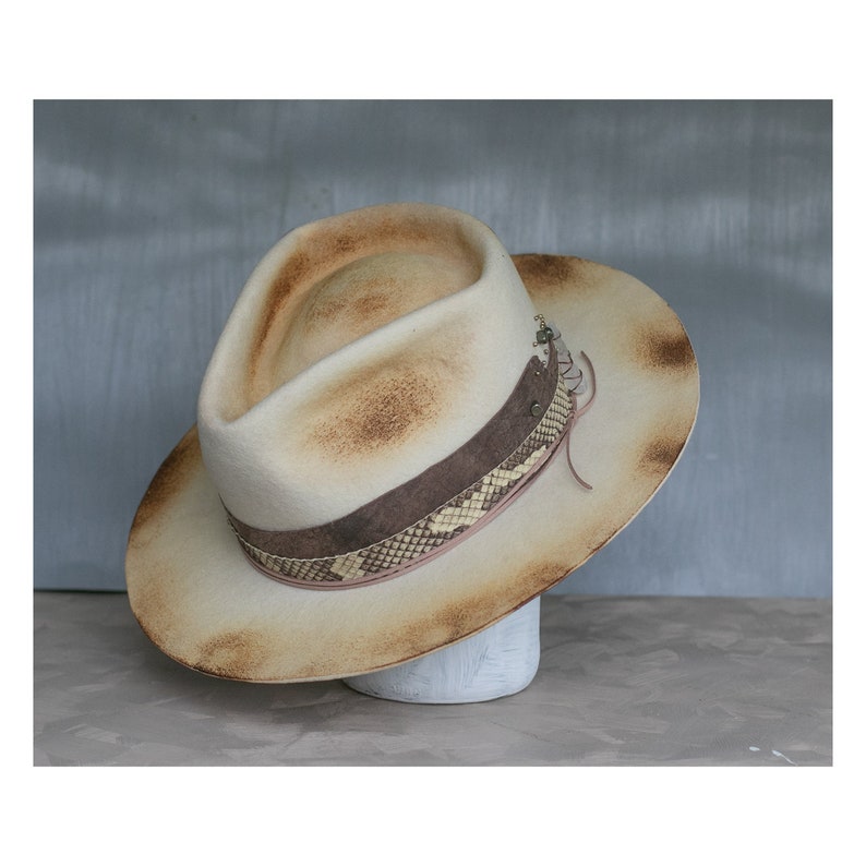 May include: A light brown felt fedora hat with a snakeskin band and a brown leather strap. The hat has a distressed look, with brown stains and a faded appearance.