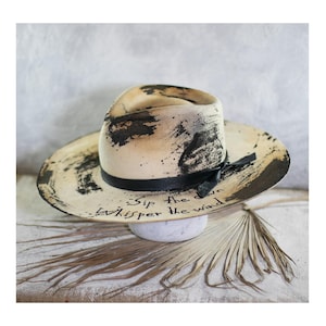 May include: A cream-coloured fedora hat featuring black paint splatters and brushstrokes. A black ribbon band encircles the crown. The brim displays the handwritten text "Sip the dawn" and "Whisper the wind" in black ink, creating a distinct contrast.