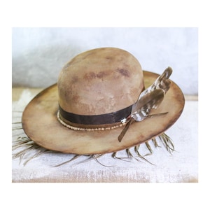 Puede incluir: Un sombrero vaquero de cuero marrón con un ala ancha y un adorno de plumas. El sombrero tiene un aspecto desgastado y una banda de cuero marrón con cuentas blancas.
