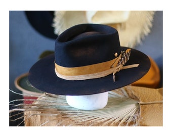 May include: A black felt fedora hat featuring a wide, stiff brim and a pinched crown. The hat is accented with a tan leather band, a small metallic stud, and a brown and white patterned feather secured by a thin, knotted leather cord.