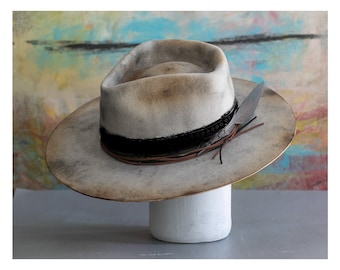 May include: A light gray felt fedora hat with a black band and brown leather accents. The hat has a distressed look and is decorated with a feather.