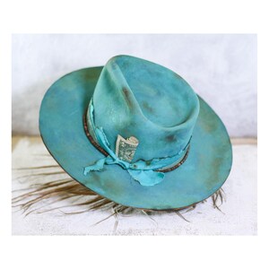 May include: A wide-brimmed fedora hat in a distressed turquoise blue finish with brown shading. The hat features a matching turquoise fabric band tied in a knot, with a rolled-up one-dollar bill tucked into the band. The brim has a raw edge.