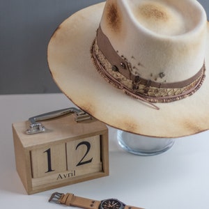 May include: A light brown felt fedora hat with a brown leather band and a decorative trim. The hat is sitting on a glass surface. A wooden calendar with the date "12 Avril" is on a white surface. A brown leather wristwatch with a black face is also on the white surface.
