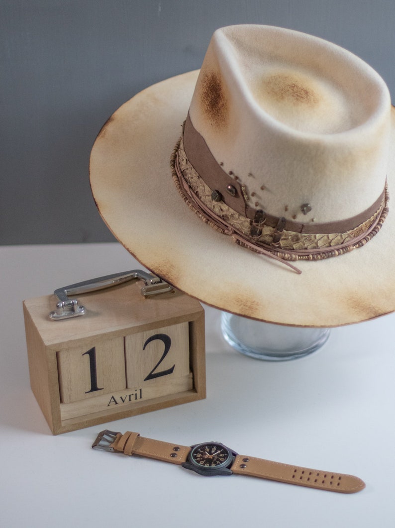 May include: A light brown felt fedora hat with a brown leather band and a decorative trim. The hat is sitting on a glass surface. A wooden calendar with the date "12 Avril" is on a white surface. A brown leather wristwatch with a black face is also on the white surface.