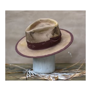 May include: A fedora hat with a distressed beige crown featuring dark brown stains and a dark maroon ribbon band. The wide brim is edged in matching dark maroon fabric, and the crown displays visible decorative stitching along the base.