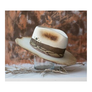 May include: A white felt fedora hat with a brown leather band and a snakeskin pattern. The hat has gold studs and a distressed look.