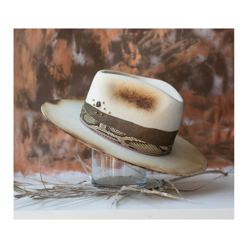 May include: A white felt fedora hat with a brown leather band and a snakeskin pattern. The hat has gold studs and a distressed look.